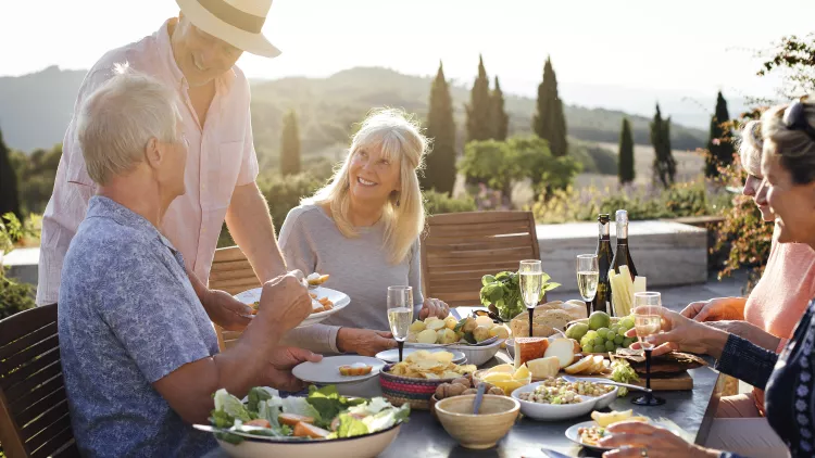 A group of mature friends are sitting around an outdoor dining table in Tuscany, Italy