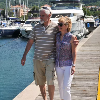 A male and female couple stood on the end of a jetty holding one another round the waist and arm with the sun on their faces.