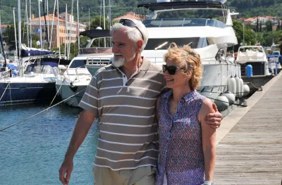 A male and female couple stood on the end of a jetty holding one another round the waist and arm with the sun on their faces.