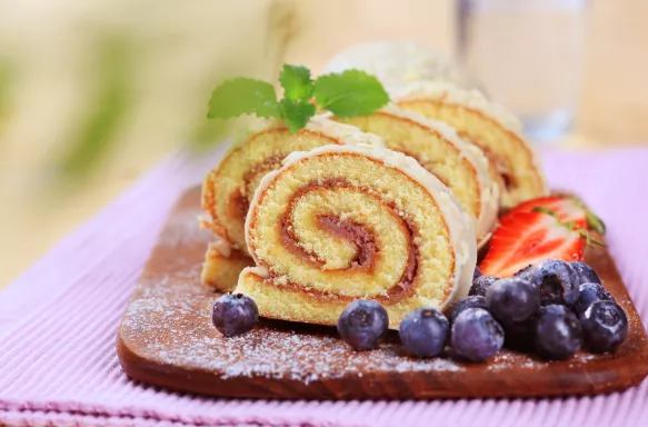 Swiss roll glazed with white chocolate icing, a side of strawberries and blueberries, lightly coated in icing sugar.