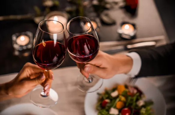 Two hands holding filled wine glasses, raised in a toast to each other above a set restaurant table