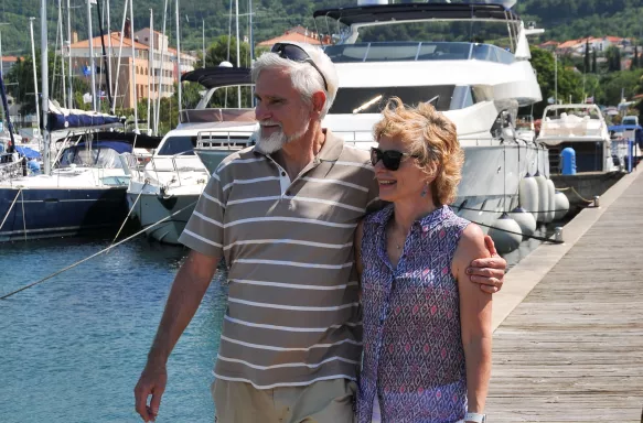 A male and female couple stood on the end of a jetty holding one another round the waist and arm with the sun on their faces.