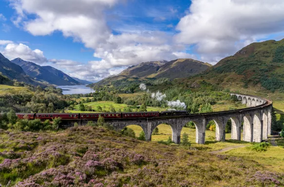 Famous Glenfinnan Railway Train amongst hills, vegetation and a lake in the background, located in Scotland.