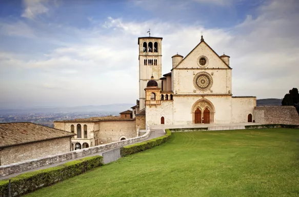 The beautiful Basilica of St. Francis of Assisi located in the town of Assisi, Italy. Photo contains a Tuscan hillside view in the background and a bright blue sky with pretty cloud formations.